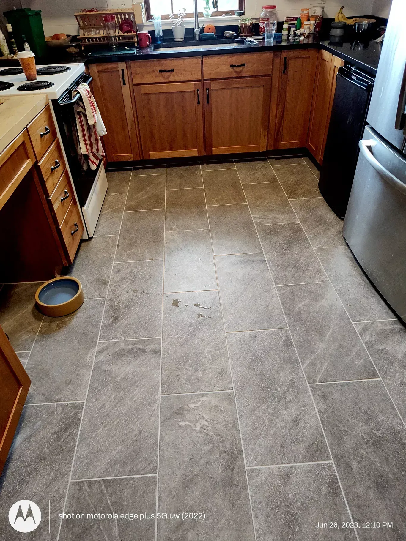 A kitchen with wood cabinets, dark countertops, gray stone-look rectangular floor tiles, and a metal refrigerator.