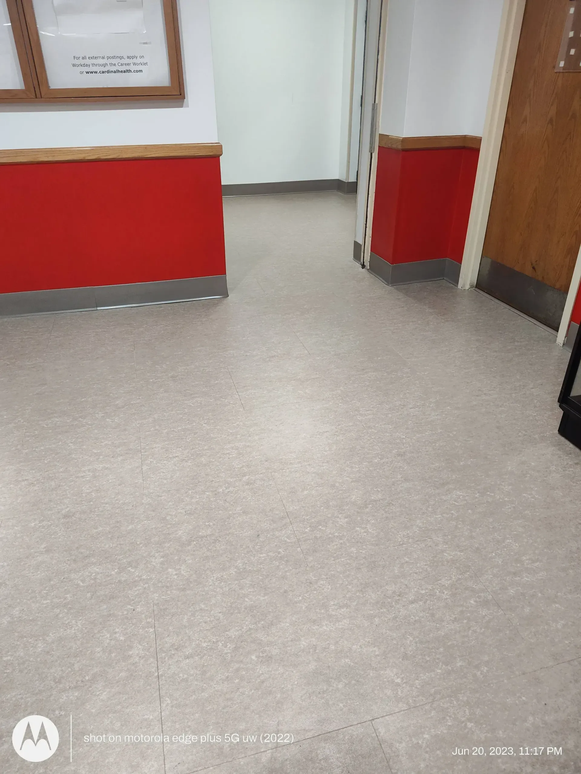 A hallway with gray speckled flooring, red walls with wooden trim, and an open doorway leading to a white interior room.