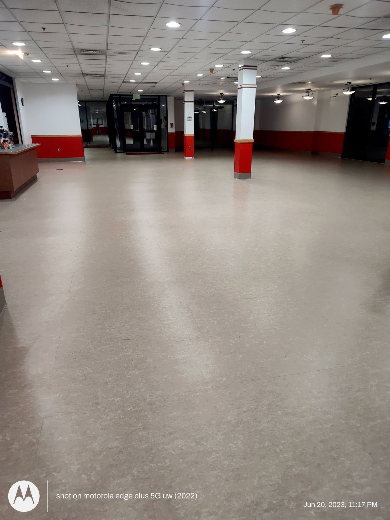 An empty commercial space with light-colored flooring, white support columns, and red wall trim under a drop ceiling.