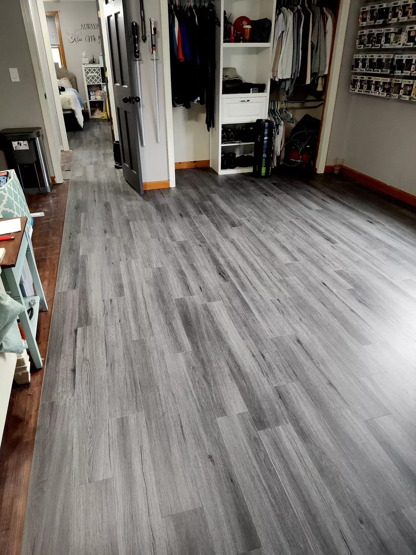 A bedroom featuring grey wood-look plank flooring, a closet with hanging clothes, and a doorway leading to another room.