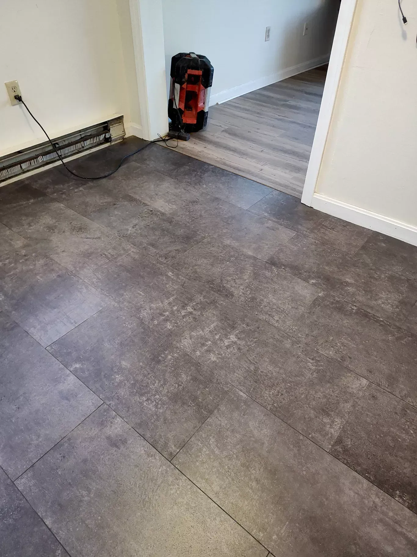 Dark gray stone-look flooring in a room transition, featuring a partially installed floor and a corner heater unit.