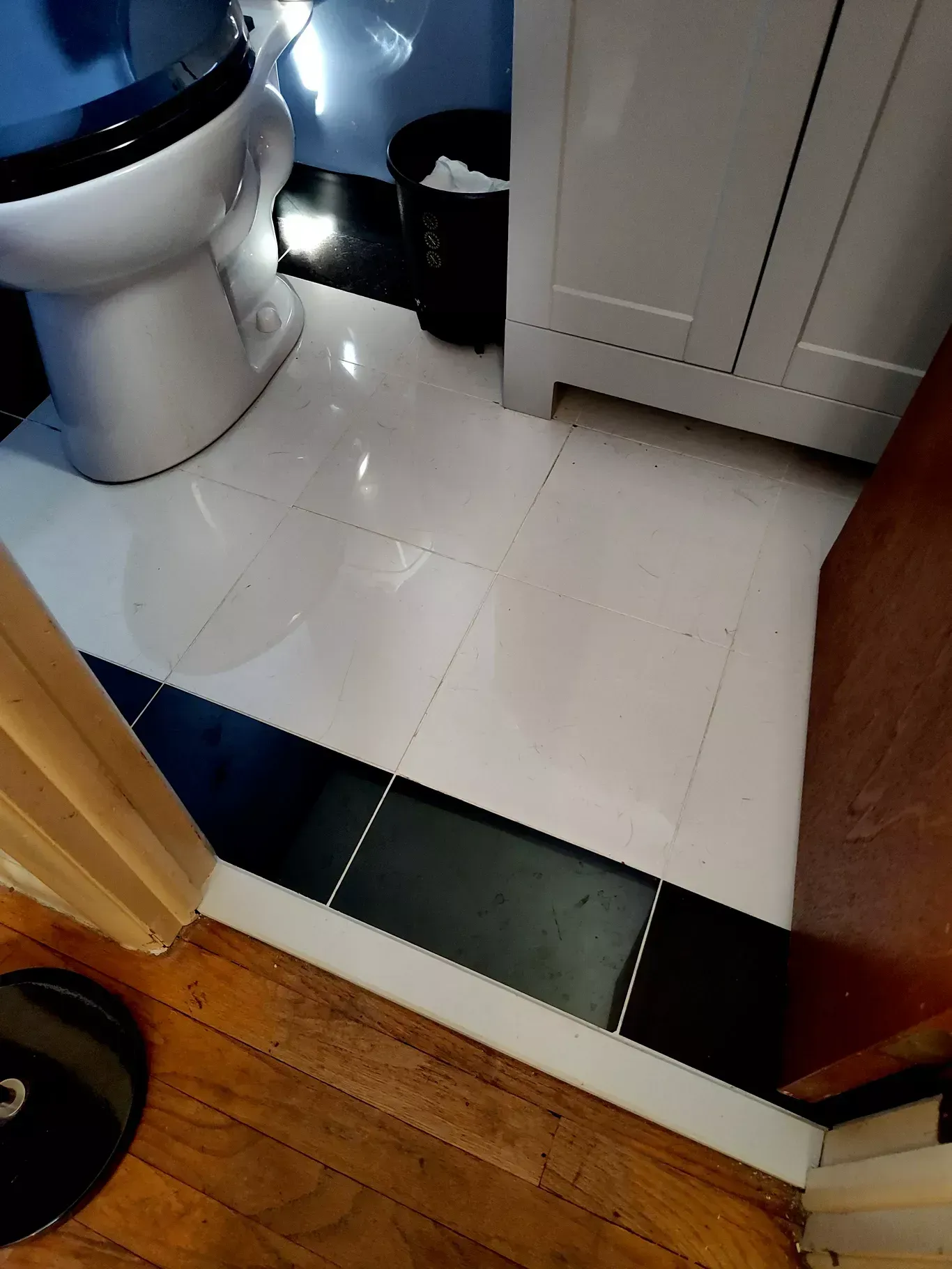 A bathroom floor with a transition from wood to white tiles, featuring a black tiled border, a toilet, and a vanity.