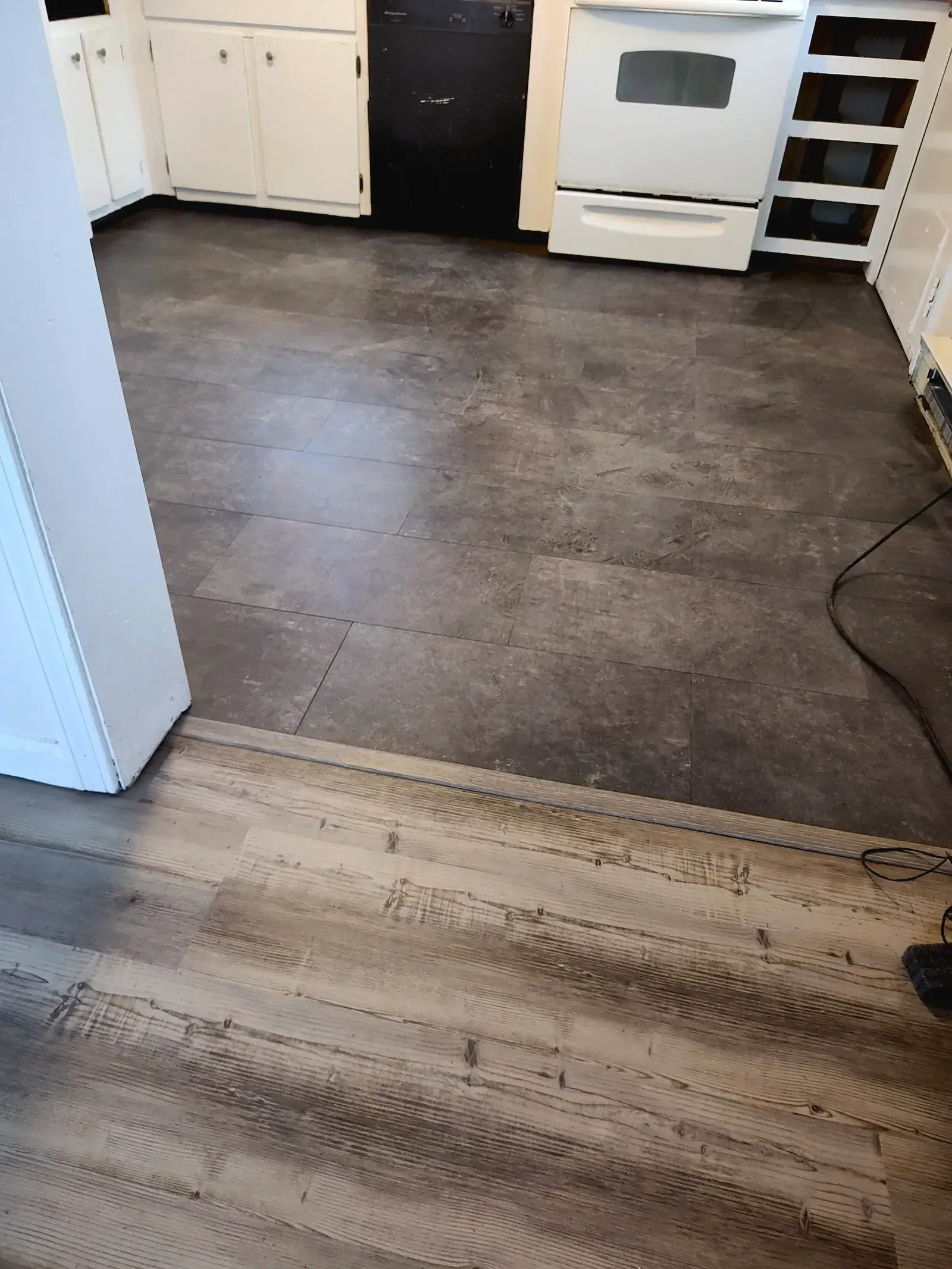 A kitchen with gray slate-look floor tiles bordering light-colored wood-look vinyl planks, white cabinets, and an oven.
