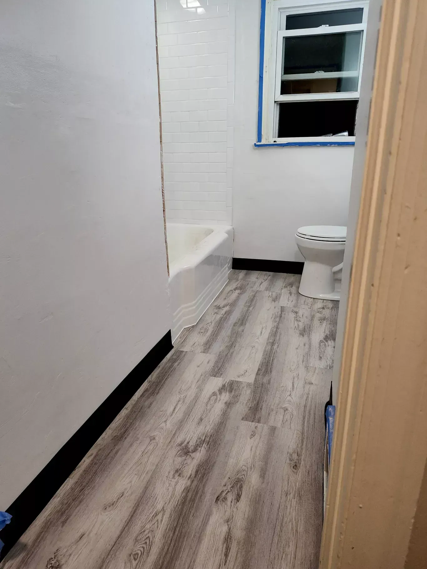 A bathroom with white walls, a bathtub with white tile surround, a white toilet, and gray wood-look laminate flooring.