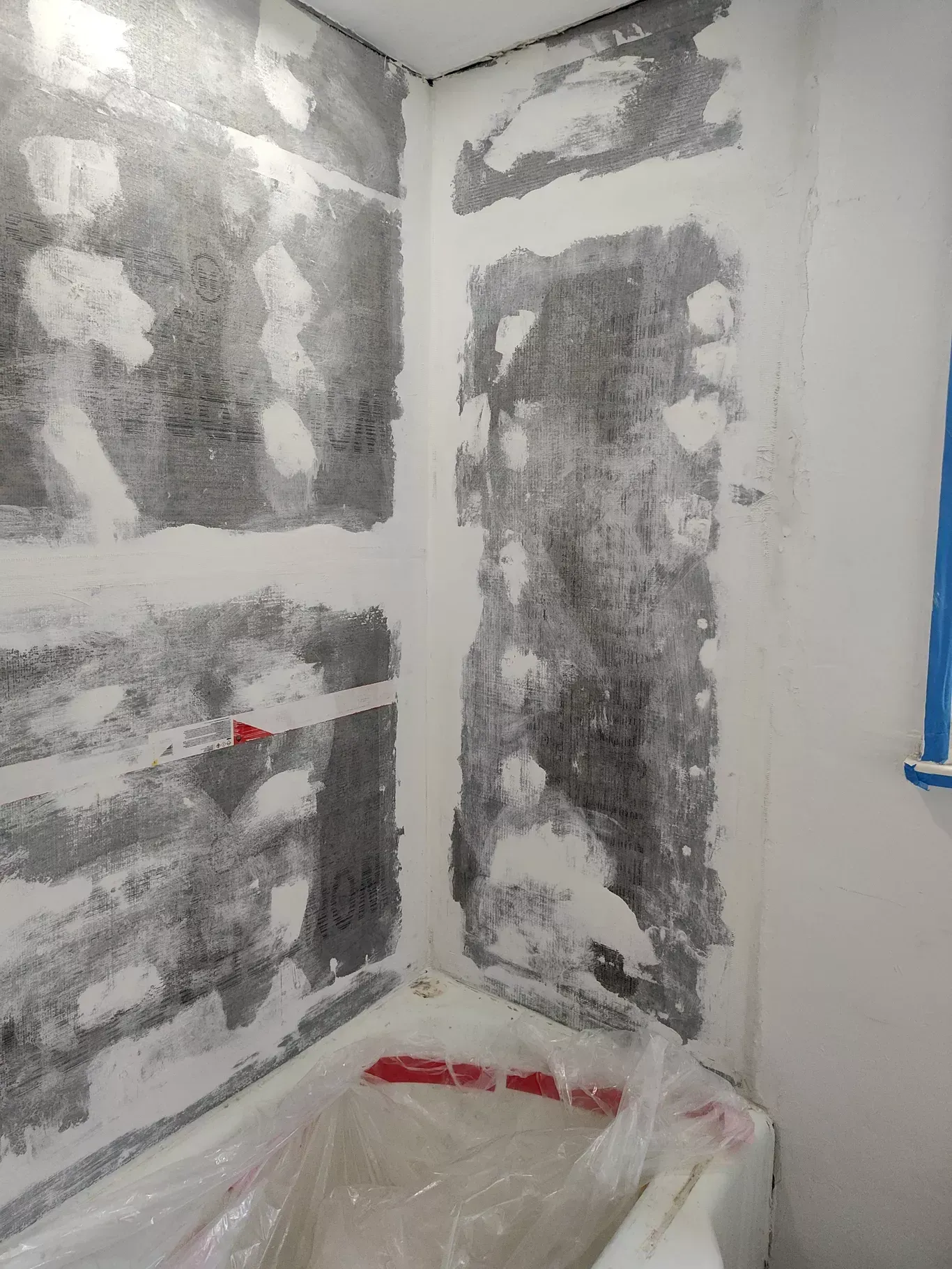 A partially plastered corner wall of a bathroom above a bathtub protected by plastic sheeting.