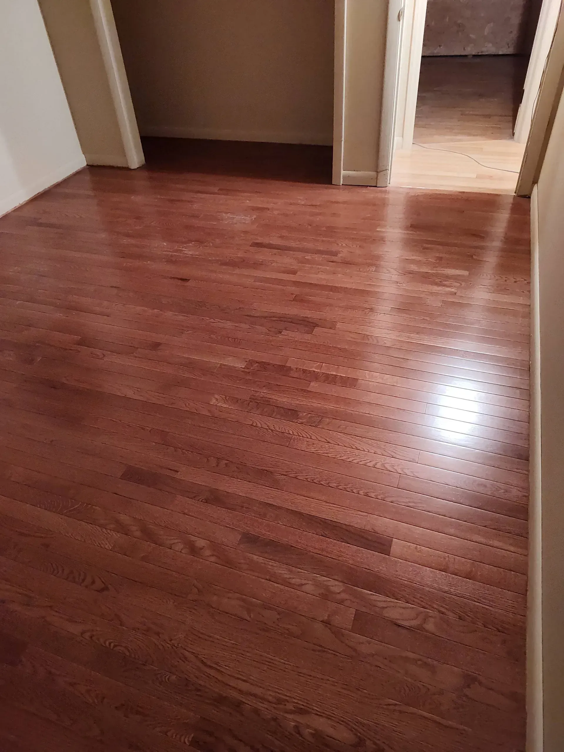 A wide shot of a room with polished, warm-toned hardwood floors, a closet doorway, and an adjacent hallway entrance.