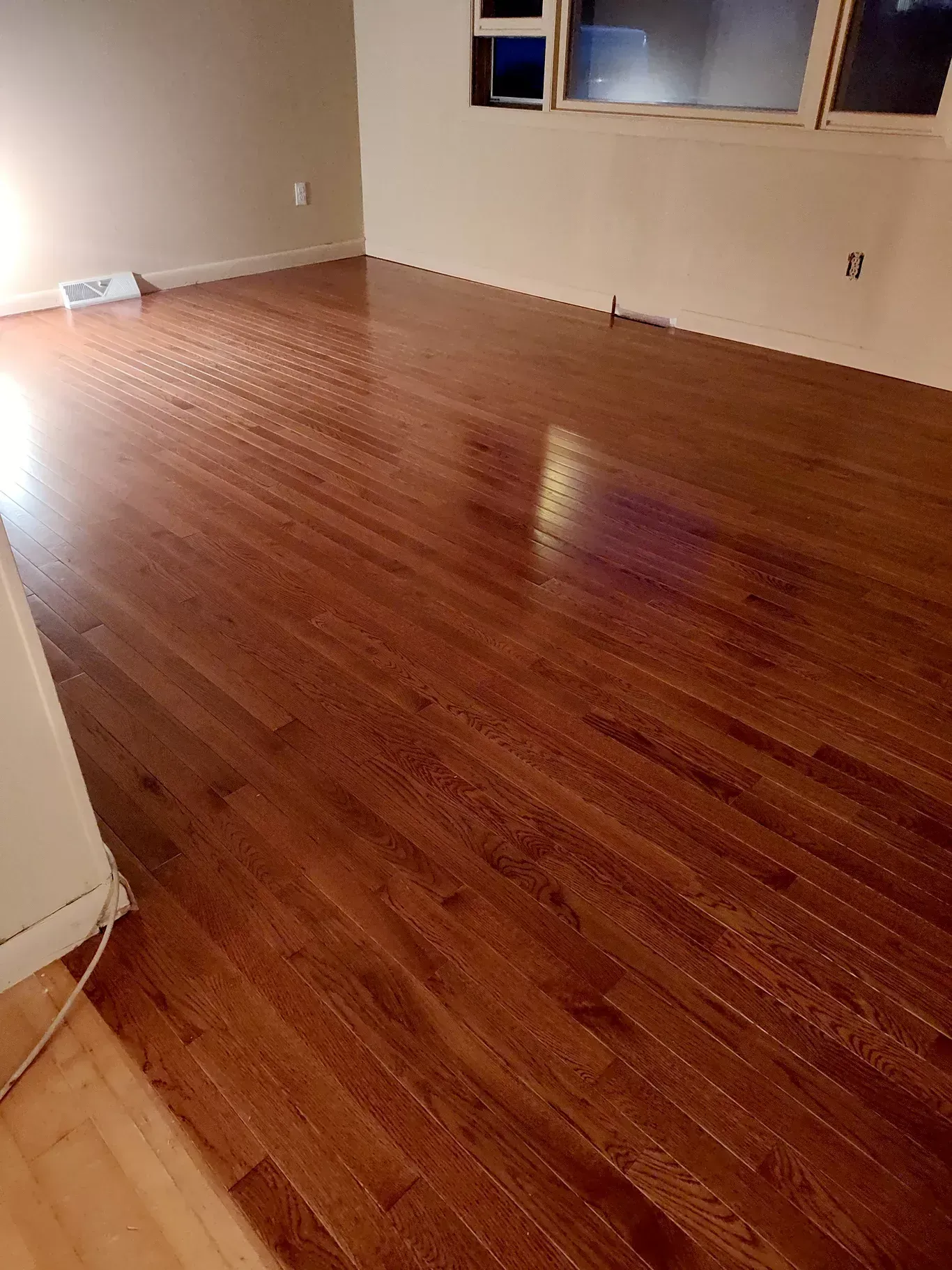 A room with polished, dark wood plank flooring, light-colored walls, and a multi-pane window.