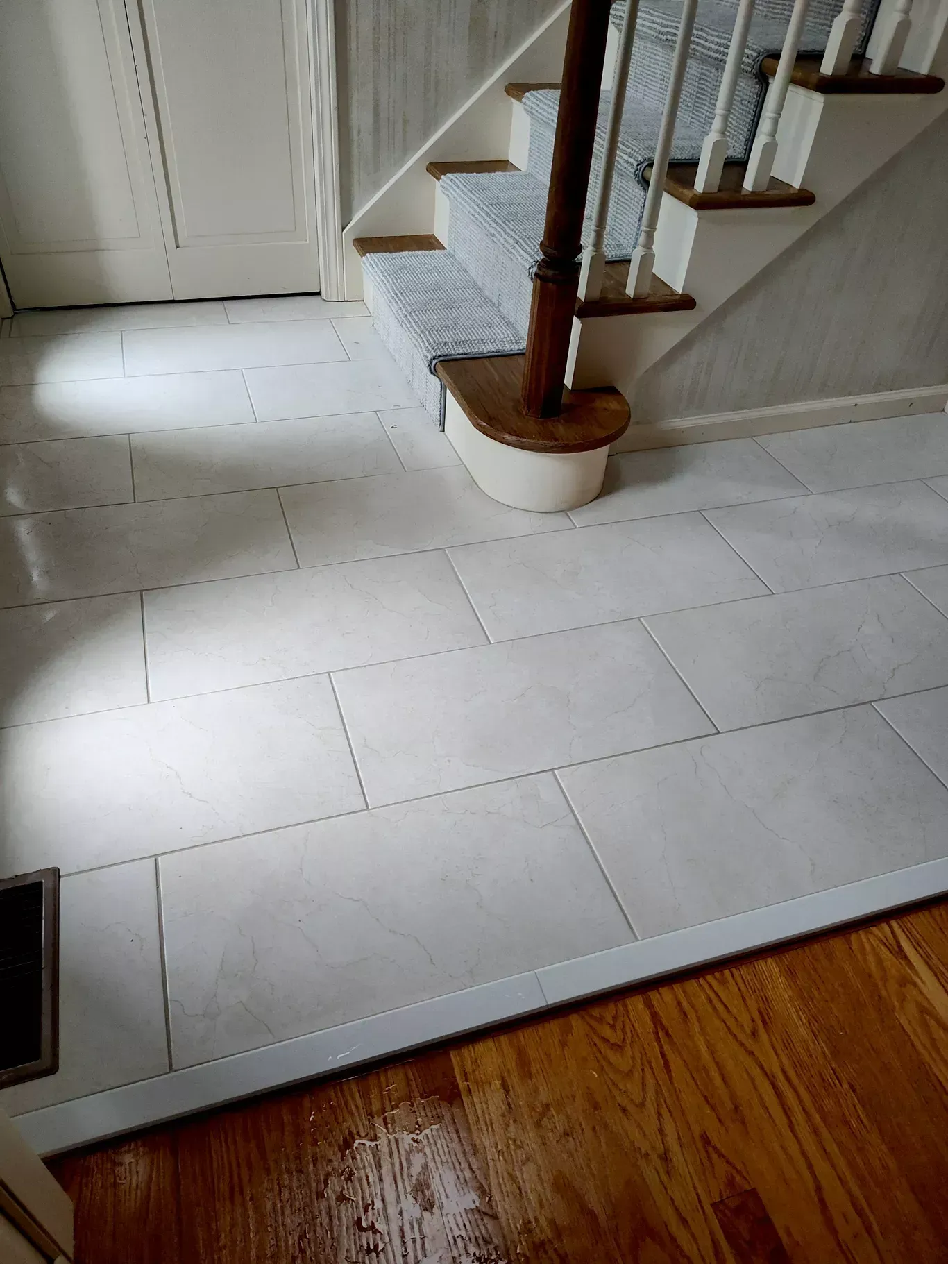 White rectangular floor tiles meeting a hardwood floor near a wooden staircase with a carpeted runner.