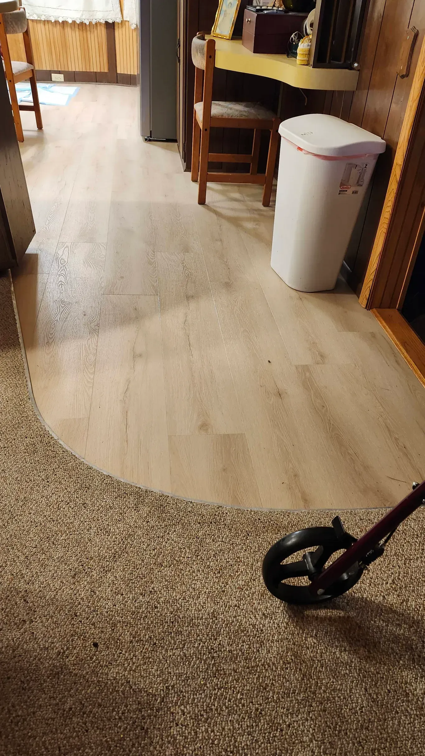 A light-colored wood floor transitions into a textured, dark carpet in a room with a white trash bin and a mobility walker.