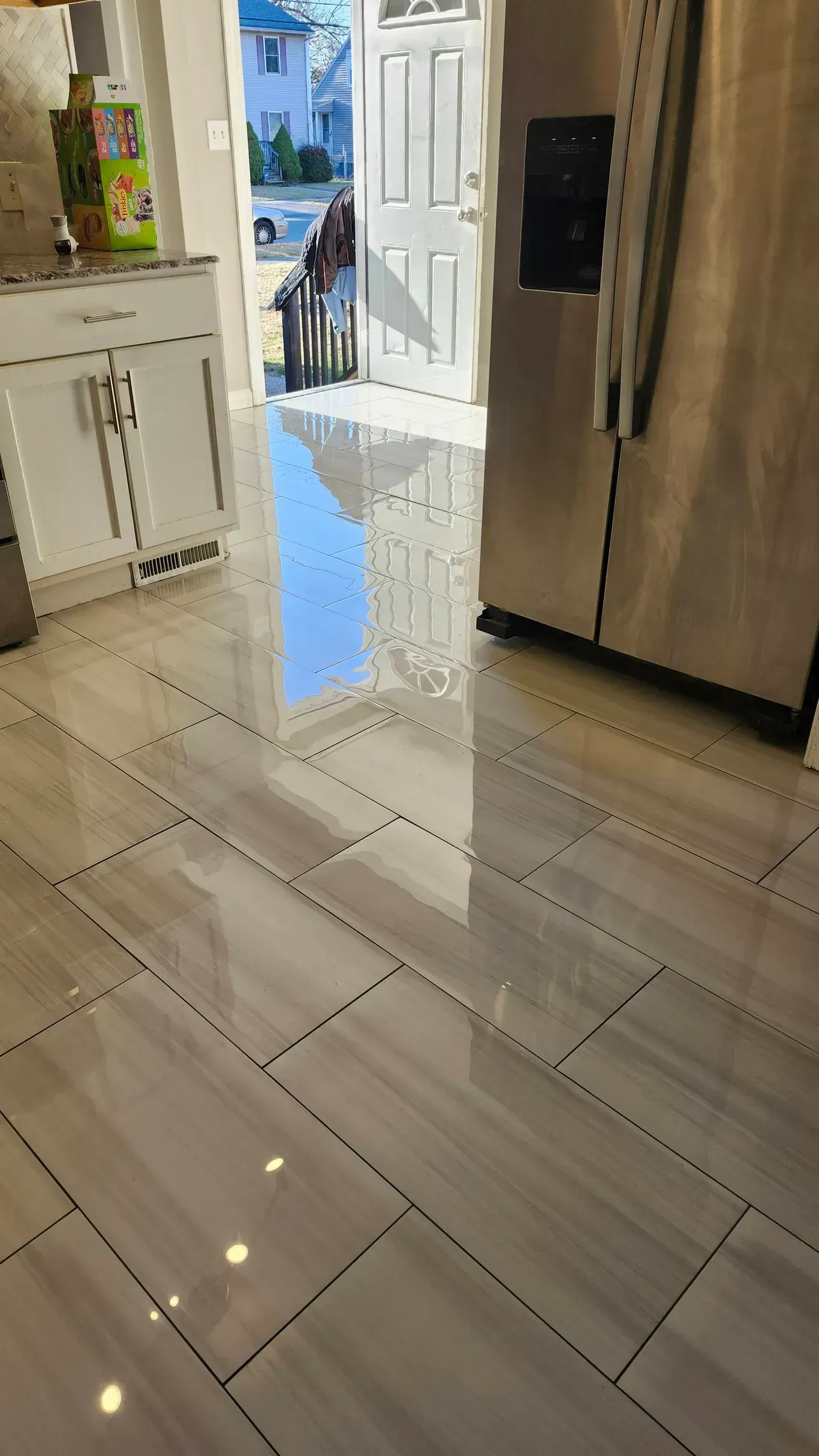 A glossy tiled floor reflects light from an open doorway in a kitchen with white cabinets and a stainless steel fridge.