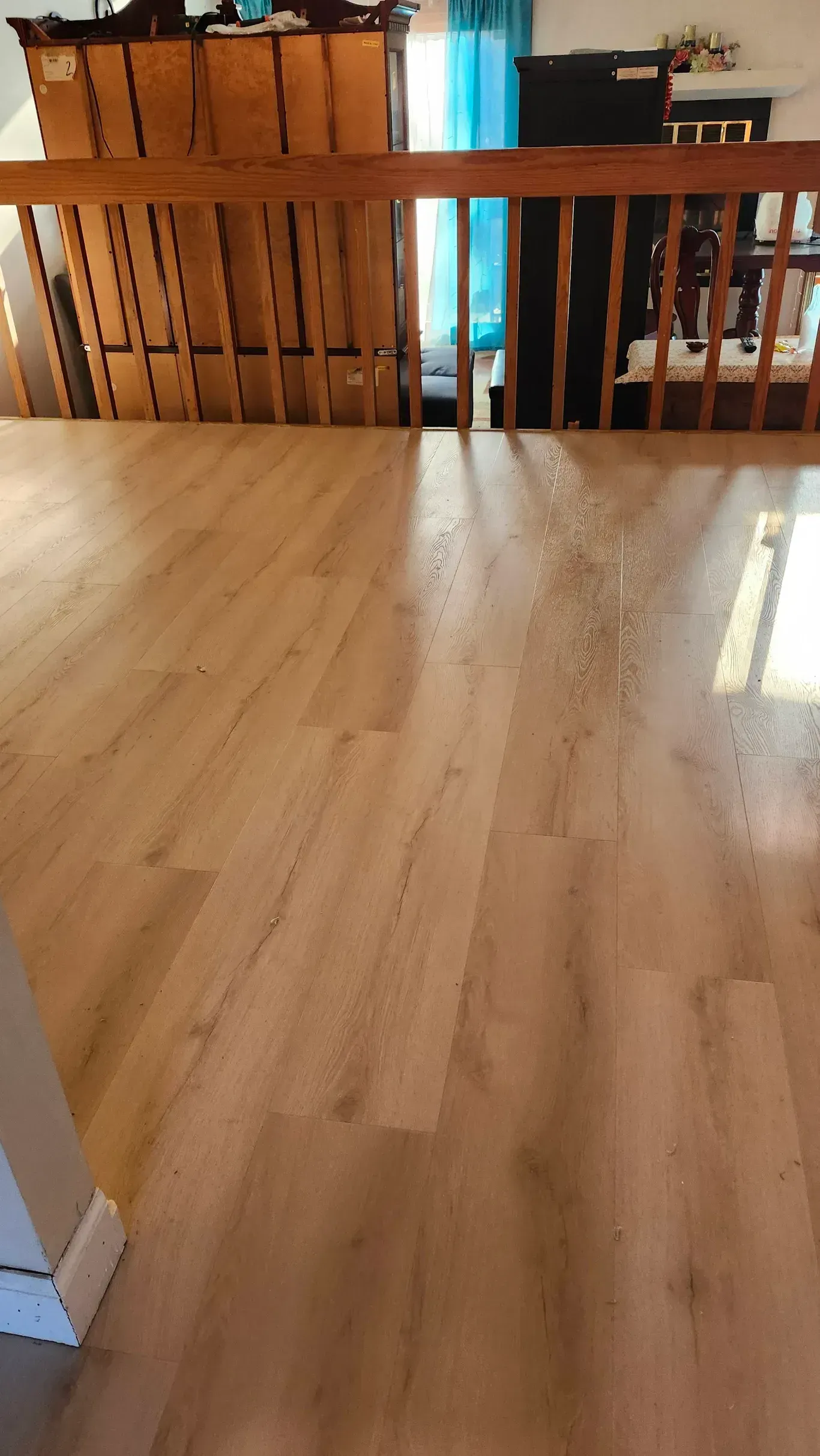 Bright sunlight streams through a wooden railing onto a light-colored wood-look laminate floor in an indoor space.