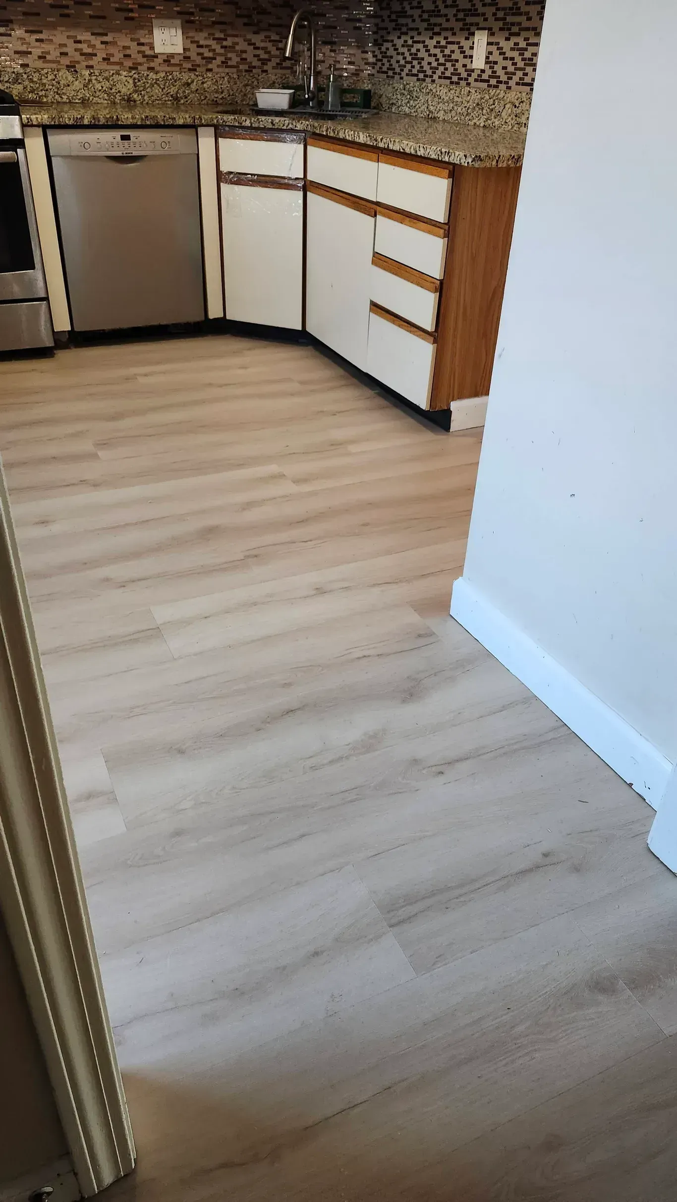 A kitchen area with light wood-look flooring, white cabinets, a stainless steel dishwasher, and mottled granite countertops.