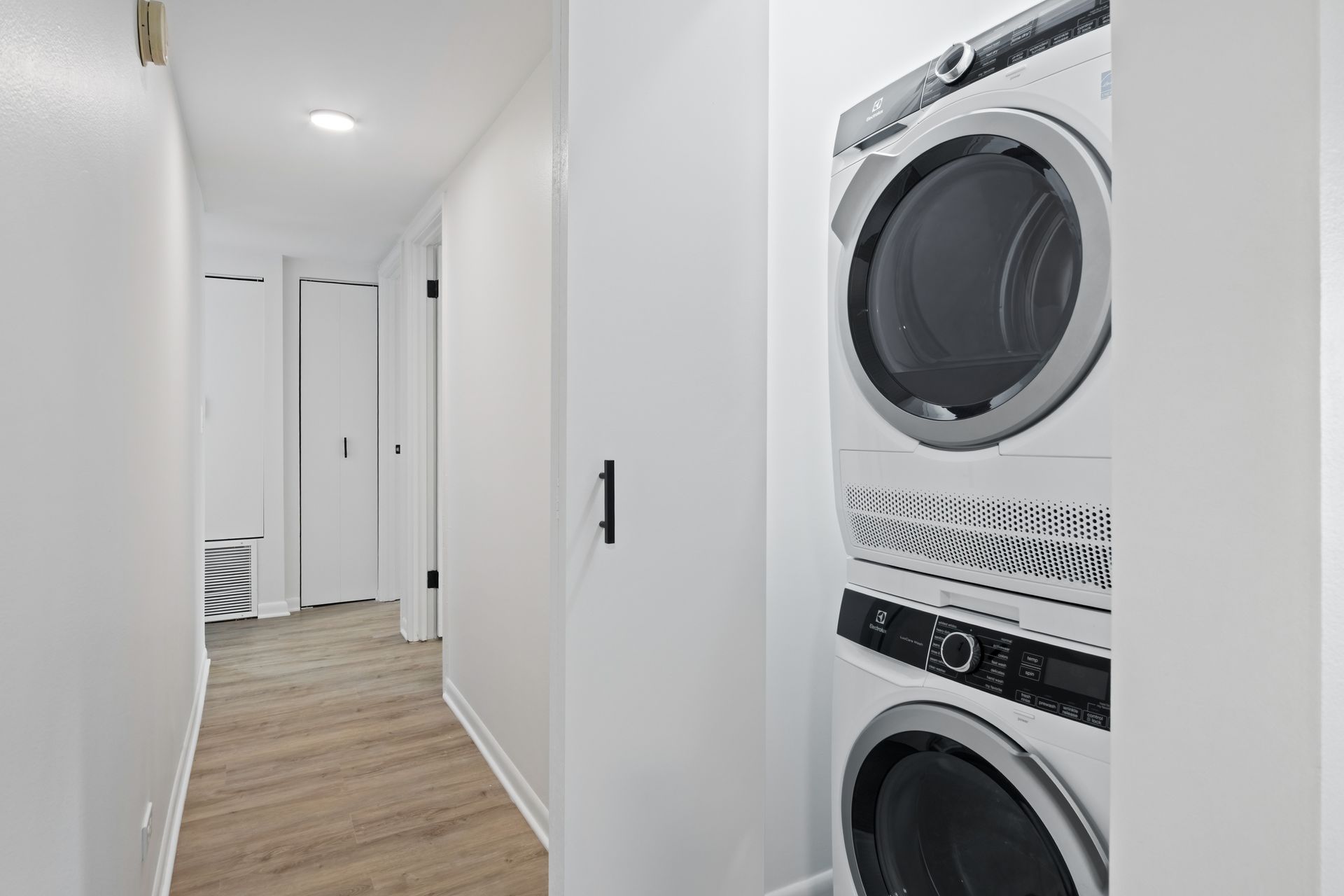 A stacked white washer and dryer in a narrow, white-walled laundry room.