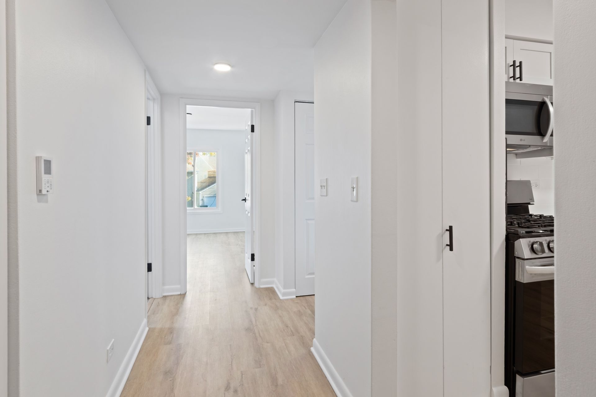 Hallway with light wood floors, leading to a room with a window; kitchen visible on the right.