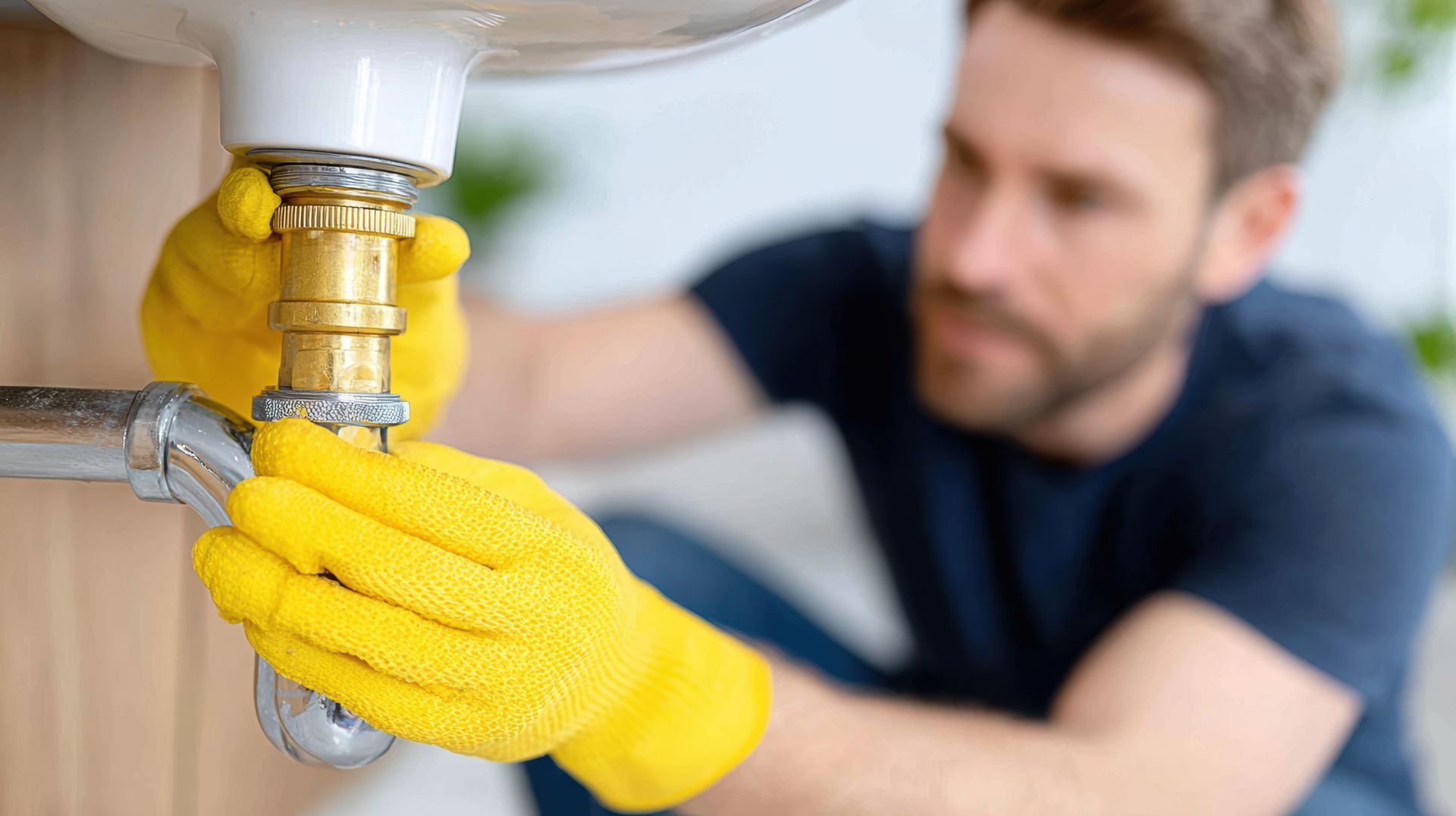 Person in yellow gloves tightening a sink pipe under a basin with a wrench, with a blurred man in the background