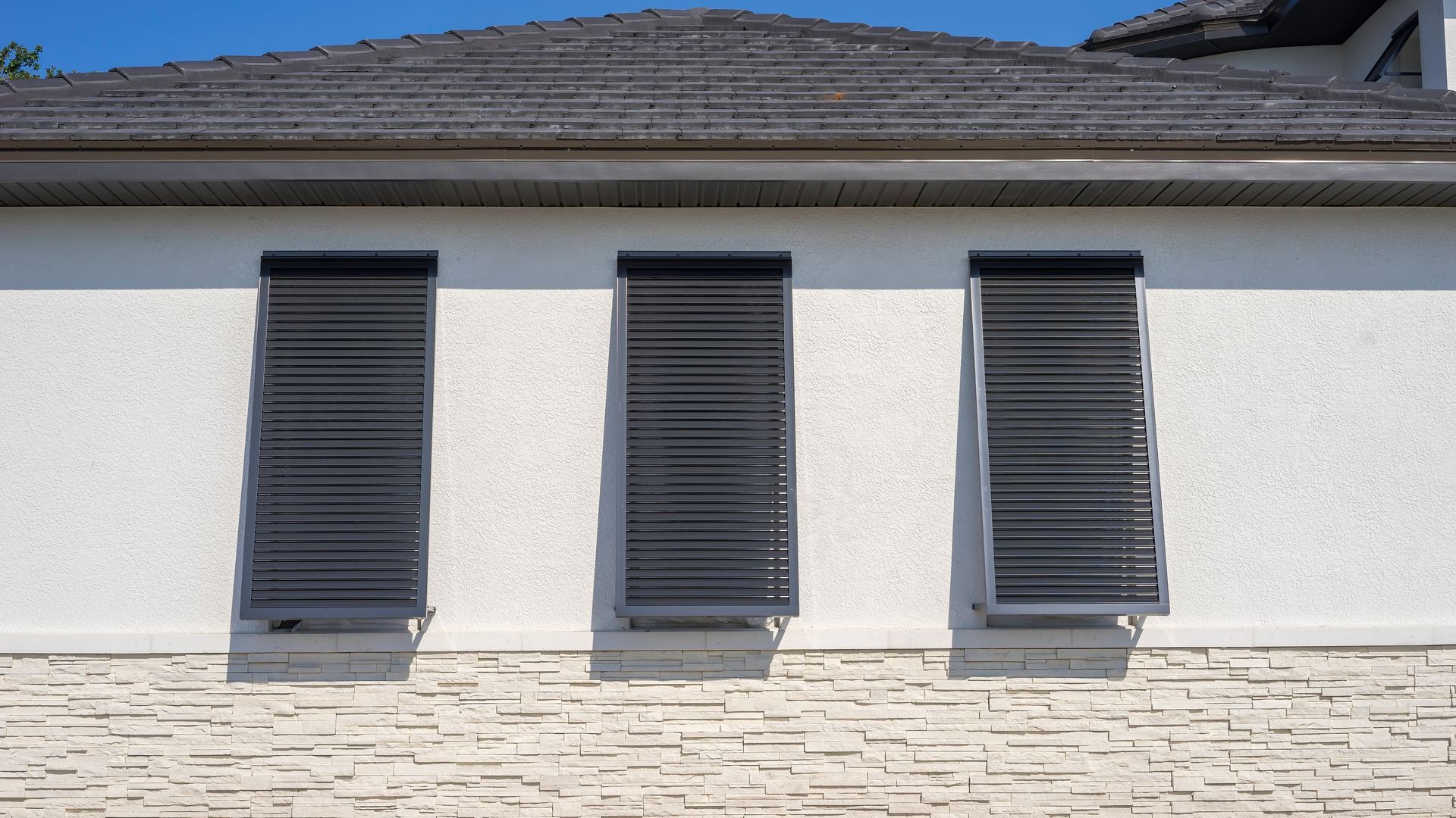 Three windows with closed, dark gray, slatted shutters on a white stucco building with a tiled roof.