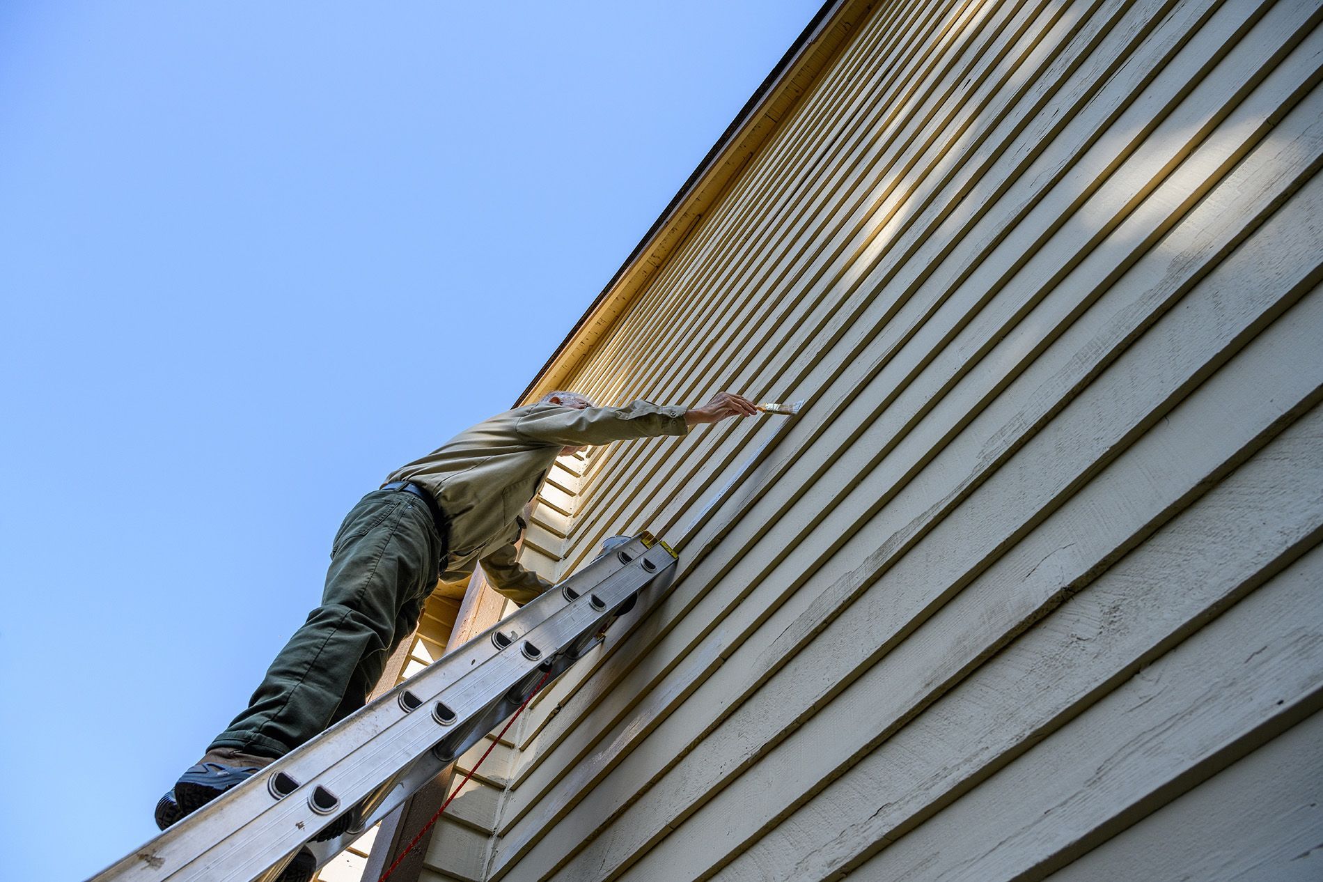 Person painting siding on a house from a ladder; blue sky in background.