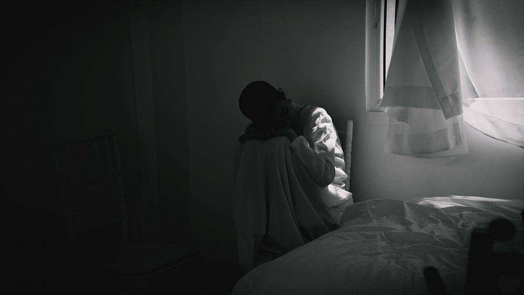 A man is sitting on the edge of a bed in a dark room.