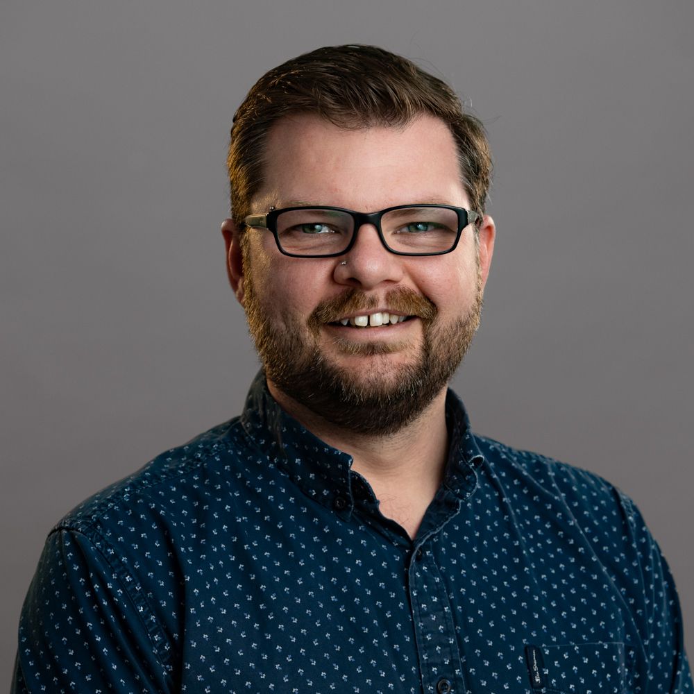 A man with a beard wearing glasses and a purple shirt is smiling.