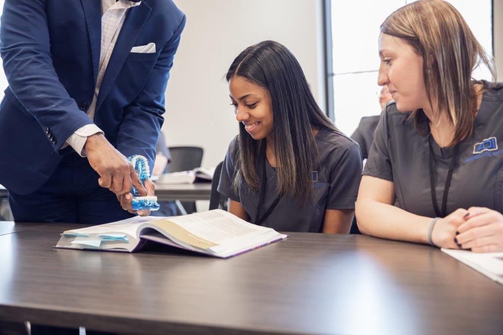 A man in a suit points at an open book with two women looking on in a classroom setting.