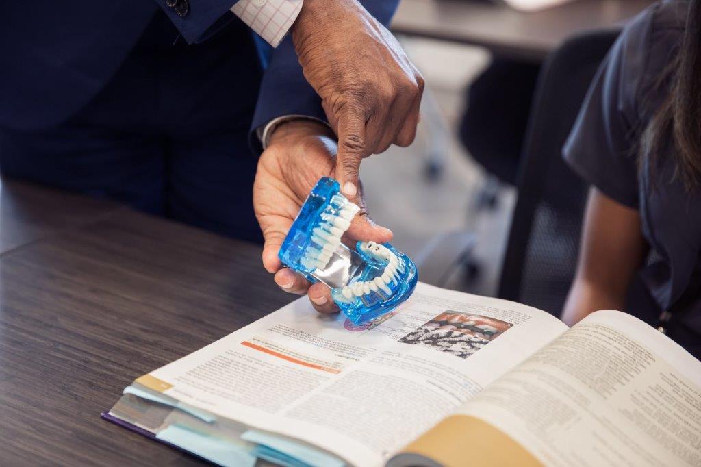 A person pointing at a blue dental mold over an open textbook, possibly in a classroom.
