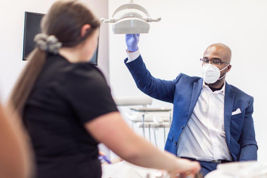 Man in a blue suit and mask adjusts dental light in a bright clinic with a woman.