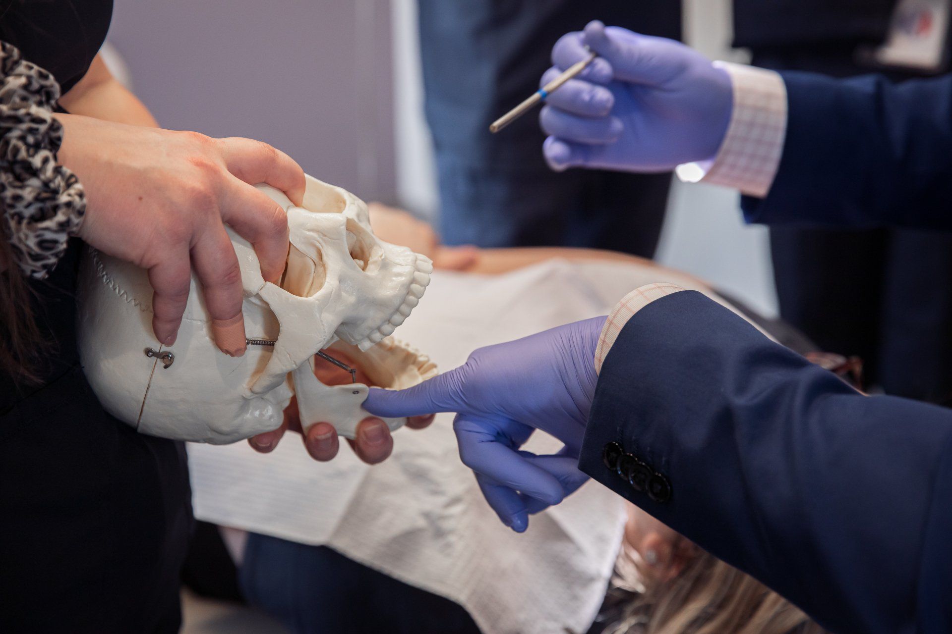 Hands holding a skull, with gloved hands pointing to the jaw. A person in the background.