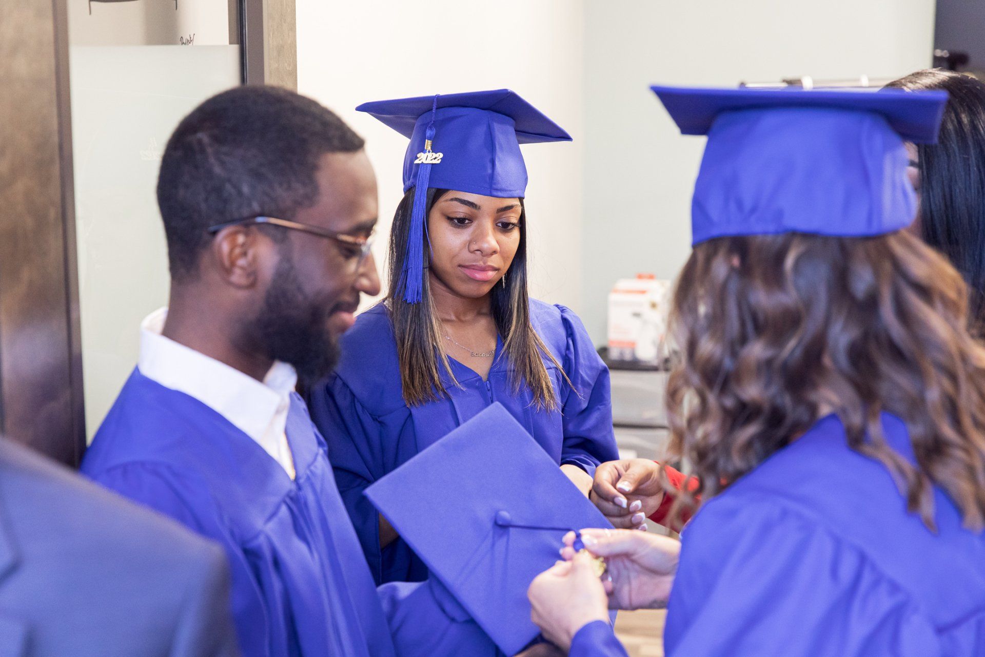 Graduates in blue robes and caps, adjusting tassels. Indoor setting, focused expressions.