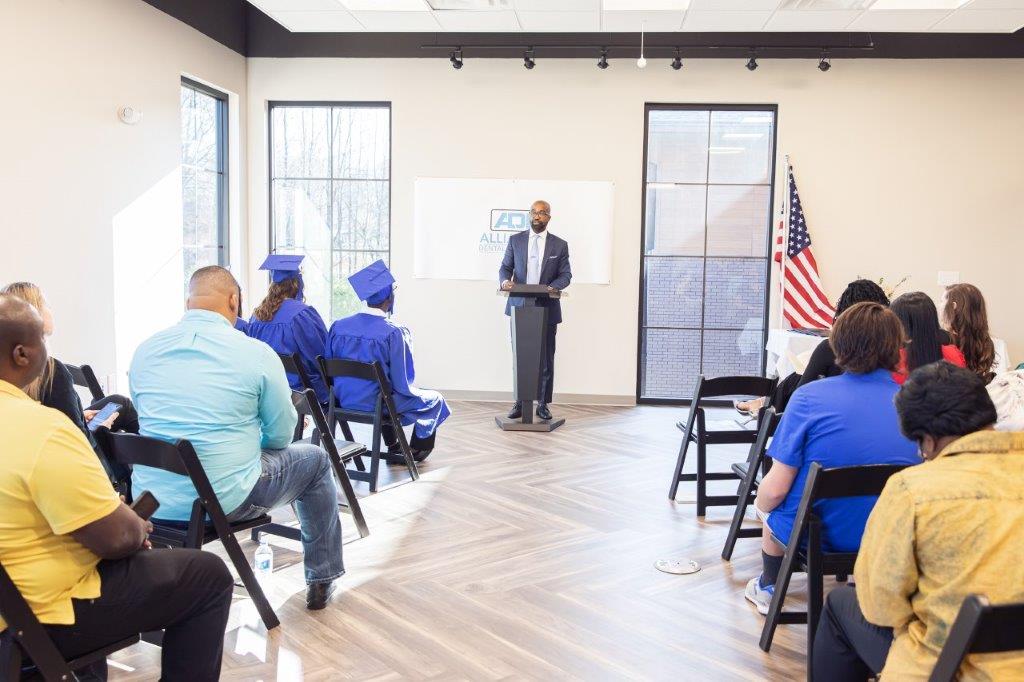 A man giving a speech in a room with people seated, including graduates in blue robes.