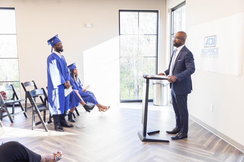 Man in suit speaks at podium. Two graduates in blue gowns and caps stand nearby. Inside a room.