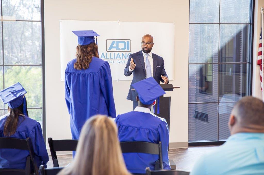 A graduation ceremony with graduates in blue gowns and a speaker at a podium in front of a banner.