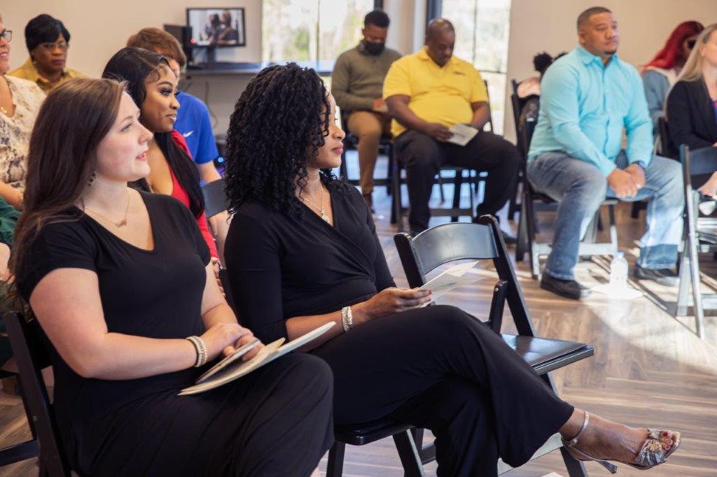 Group of people seated in a room, listening. Woman in front, in black, with notes. Others wear casual clothes.