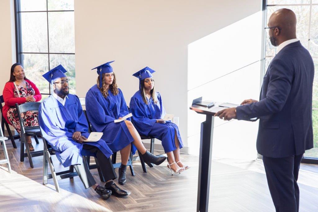 Man speaking at a podium; three graduates in blue gowns sit, listening. A woman watches. Interior, bright.