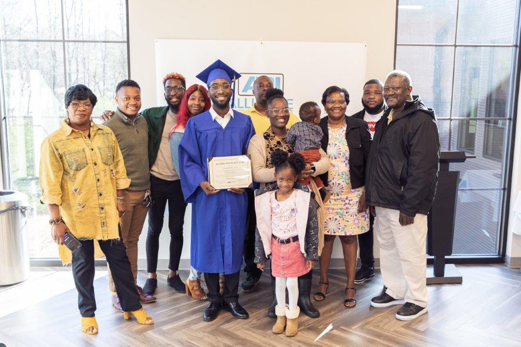 Family celebrates a graduate in blue cap and gown, holding a diploma. Setting appears to be indoors.