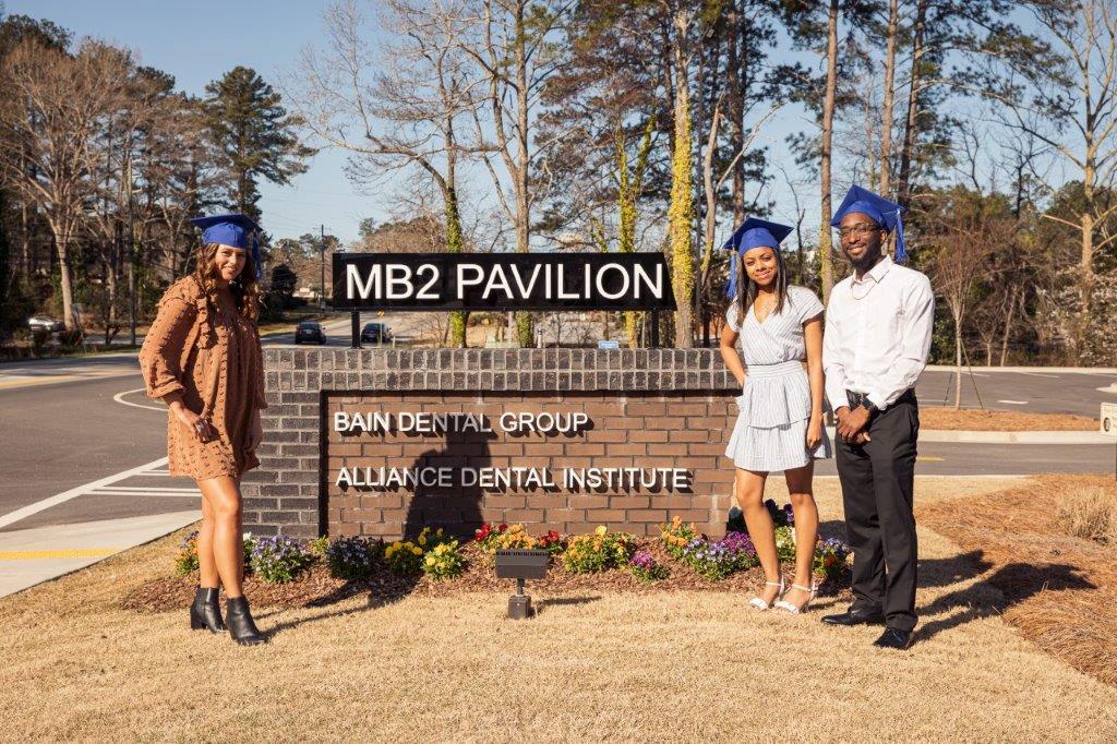 Three dental graduates in caps and gowns pose in front of the MB2 Pavilion sign.