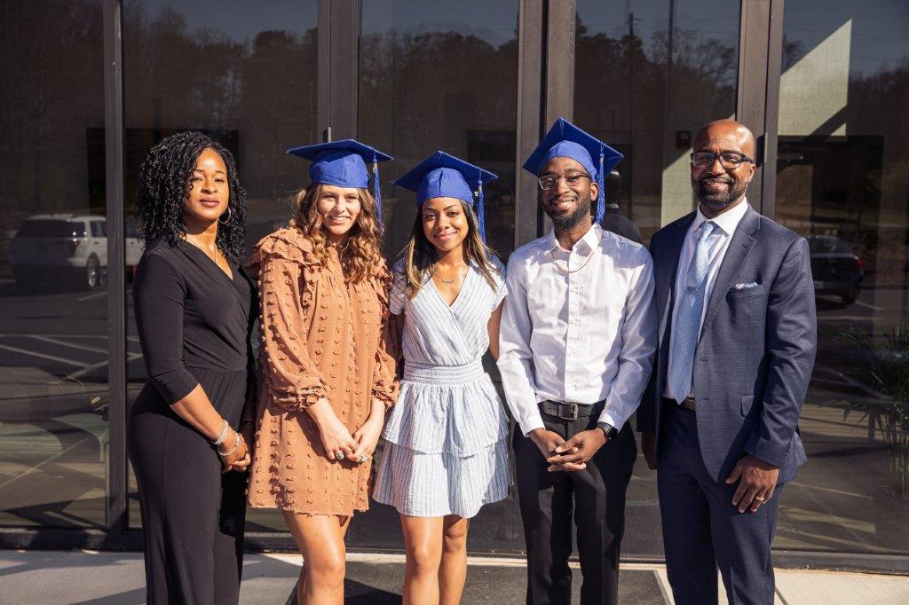 Five people pose outside: graduates in blue caps, and two adults. Sunny day.