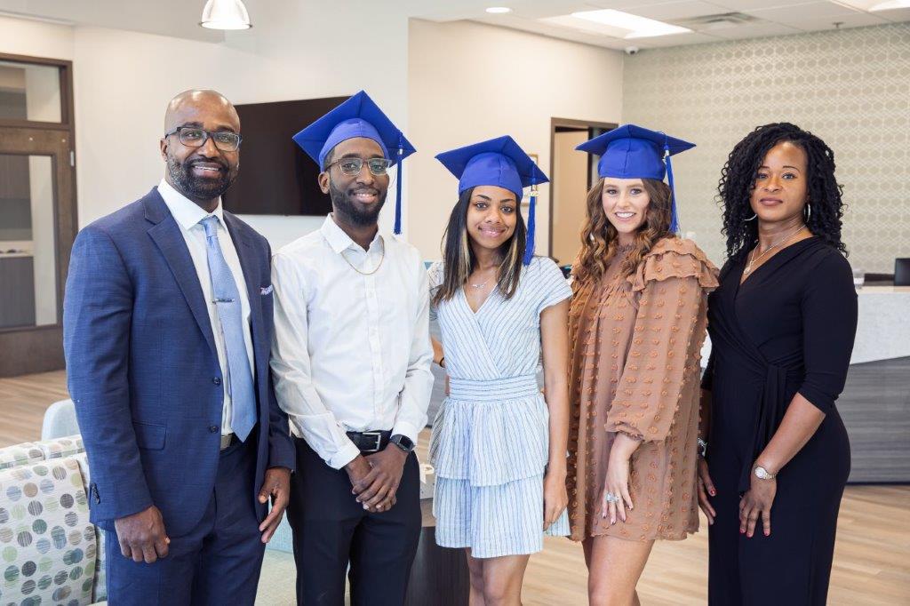 Group of five: two in suits, three in graduation caps, pose inside a well-lit modern space.