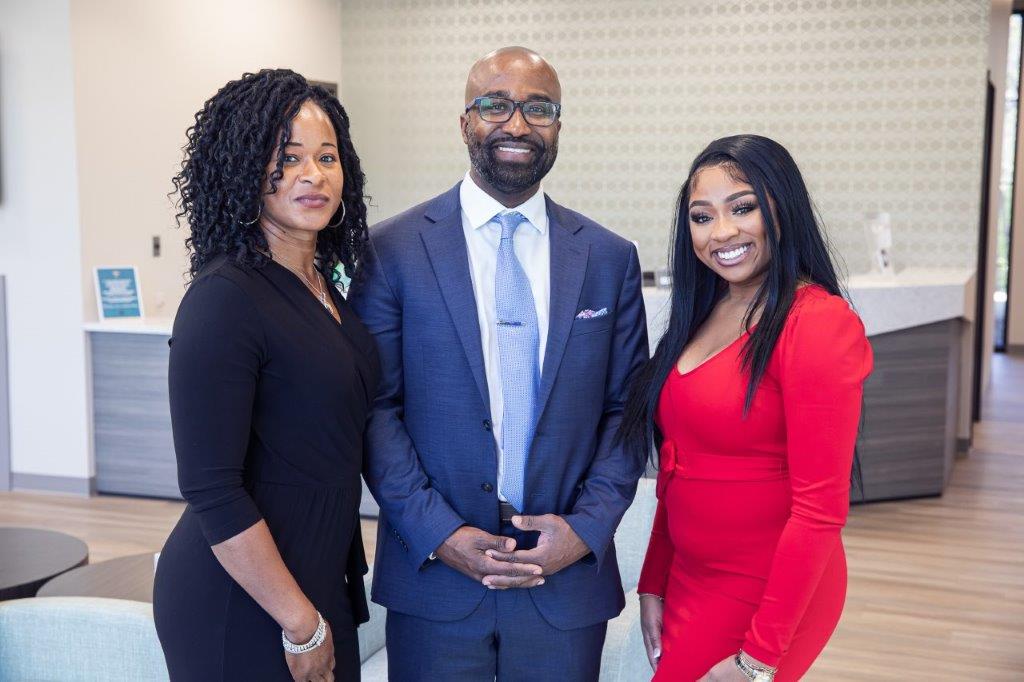 Three people posing in a modern office. Man in suit centered between two women in black and red dresses.