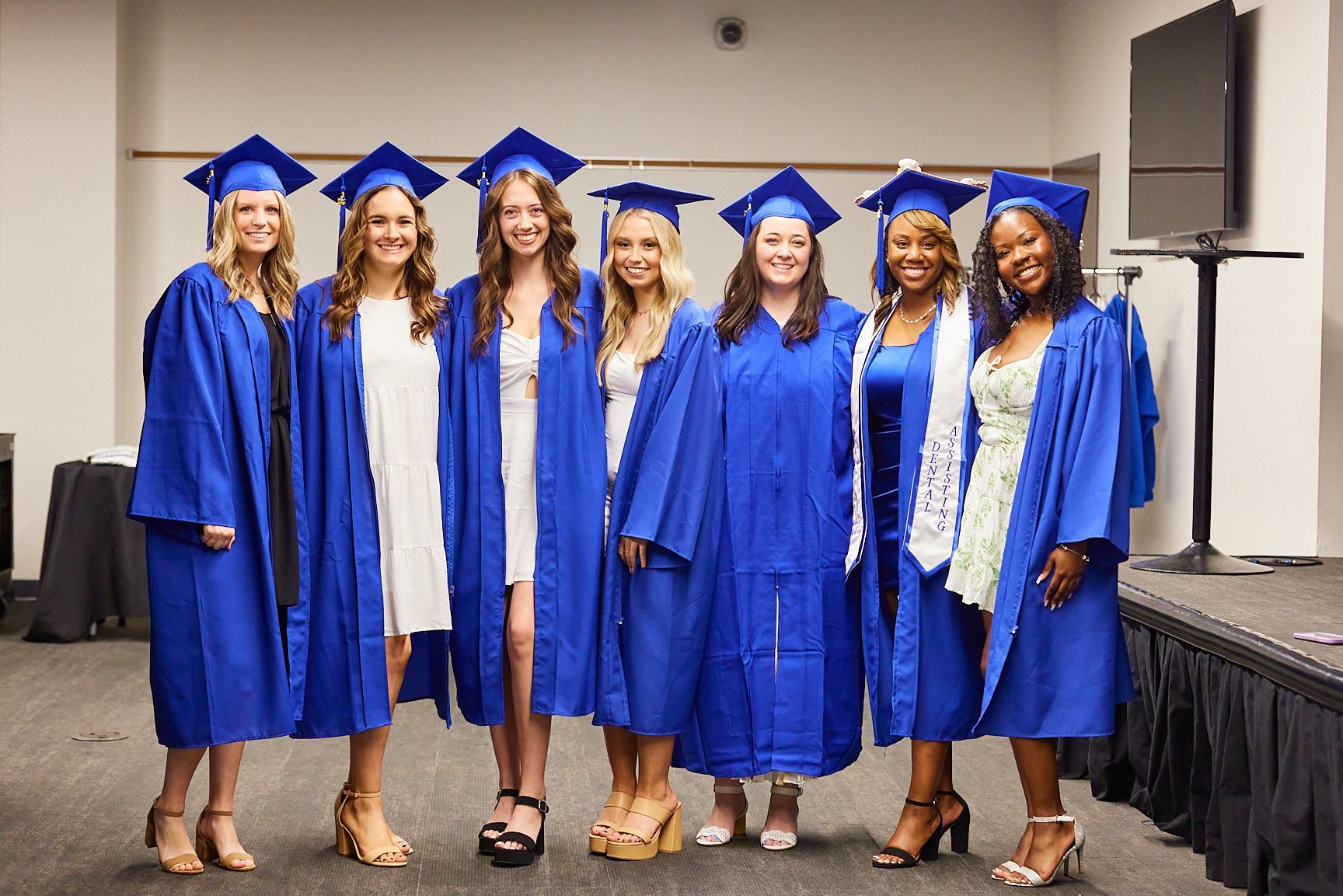 Group of seven women in blue graduation gowns and caps, smiling at a graduation event.