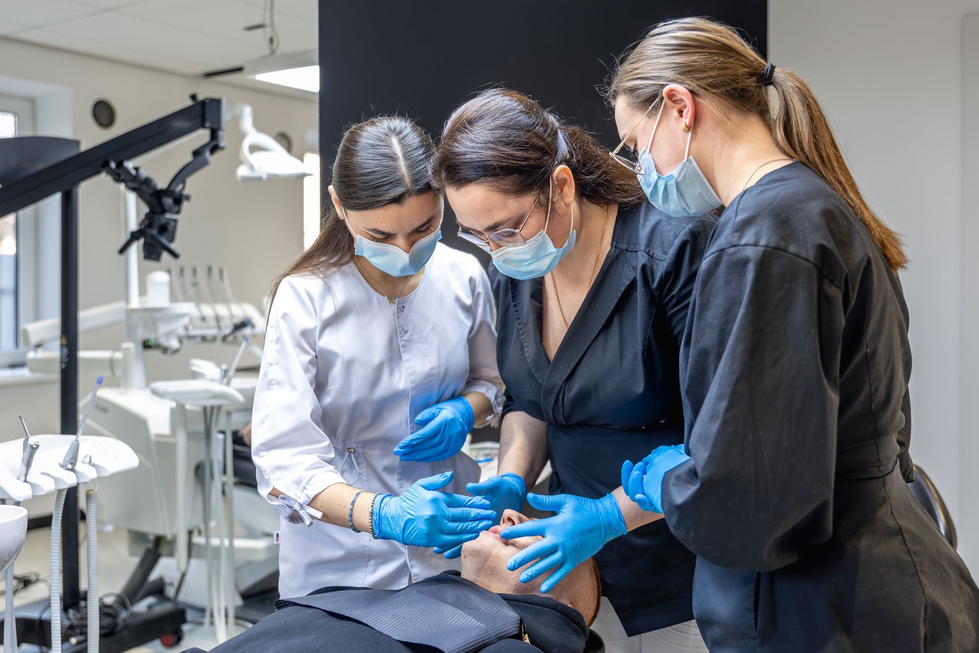Three masked clinicians in blue gloves examine a patient’s mouth in a dental clinic