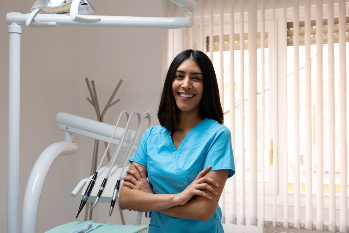 Smiling dental hygienist in blue scrubs standing in a bright dental clinic with equipment behind her