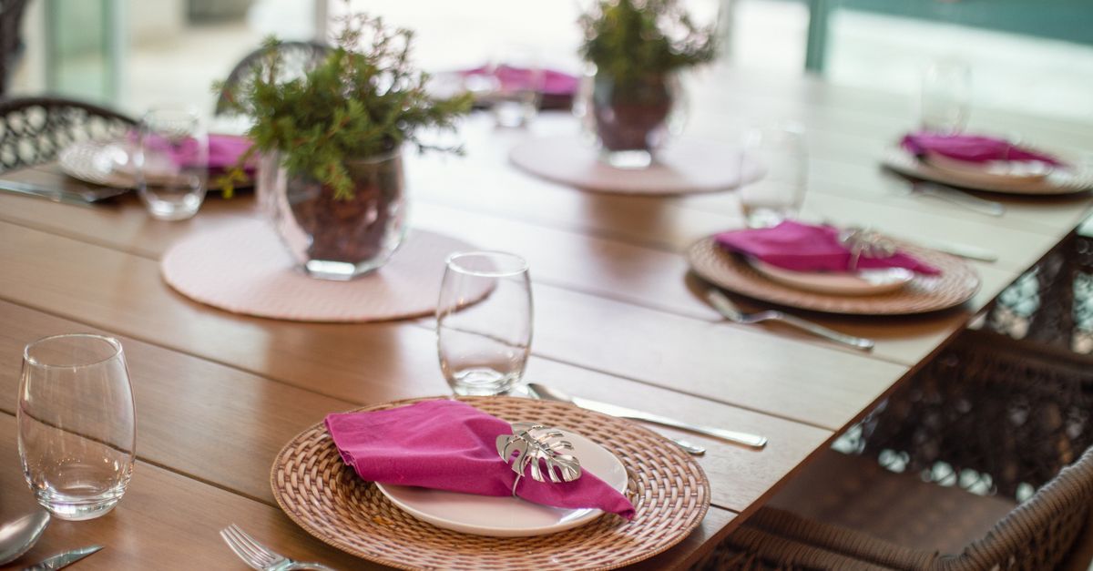 Prepara la mesa del comedor con servilletas rosas, cristalería y pequeñas plantas en macetas sobre mesas de madera.