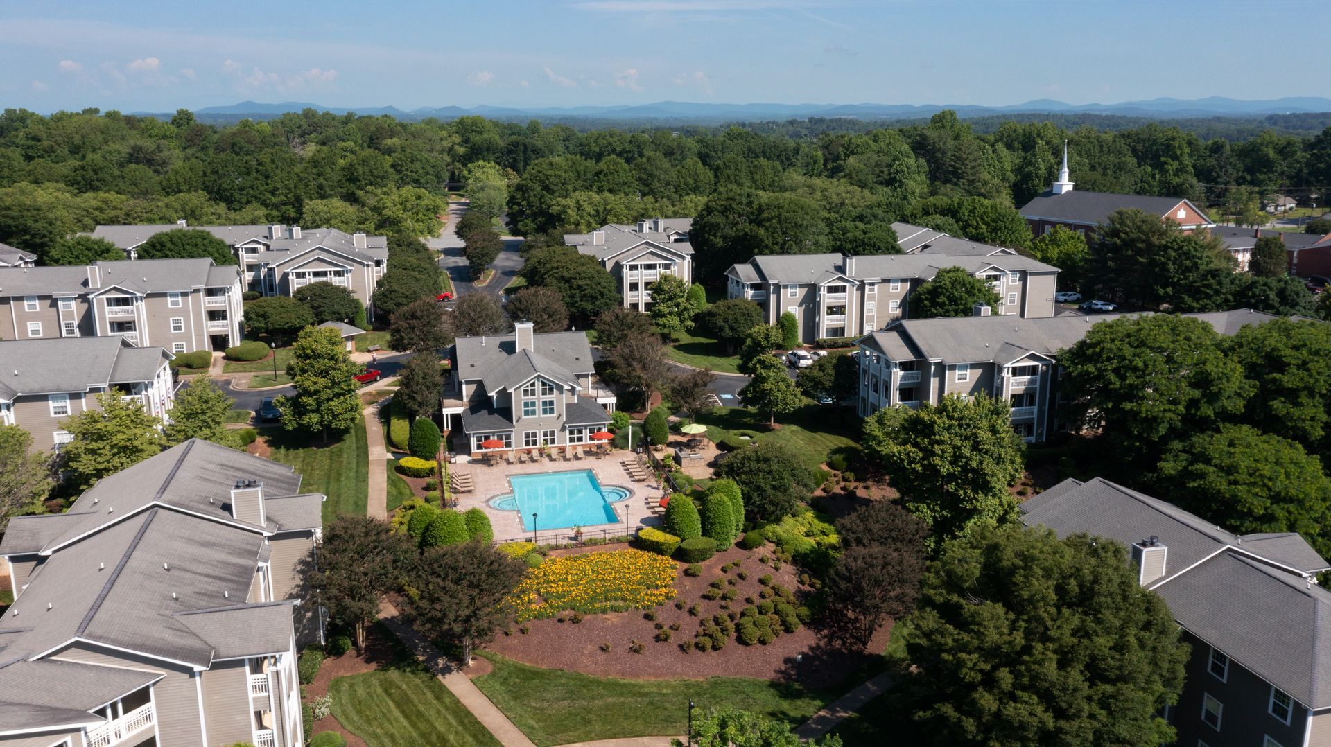 Aerial view of apartment complex with a pool, surrounded by trees, and a mountain range in the distance.