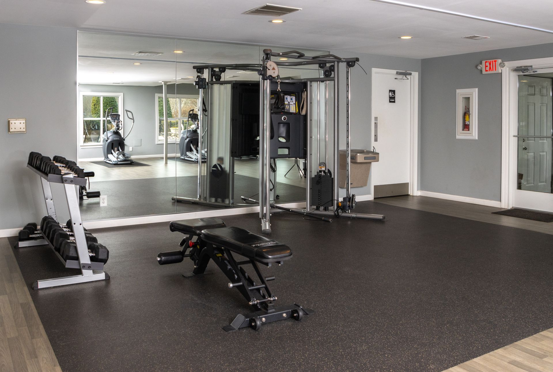 Gym with weight rack, adjustable bench, and cable machine against a mirror wall, with an exit door.