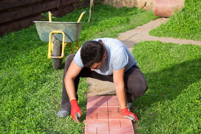 A person wearing red gloves crouches in a grassy yard, carefully placing red paving stones in a row.