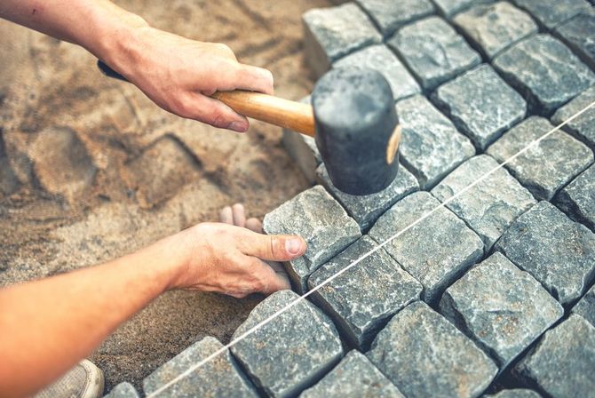 A person uses a rubber mallet to level gray cobblestones on a sand base, guided by a taut string line.
