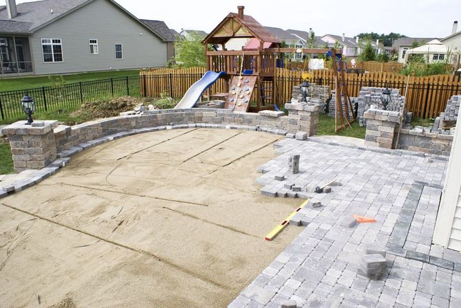 A backyard patio under construction, showing a leveled sand base, a stone seating wall, and partially laid gray pavers.