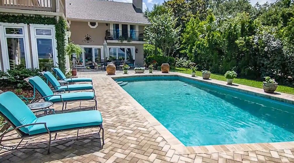 A rectangular swimming pool with blue water sits in a patio area surrounded by landscaping and a black privacy fence.