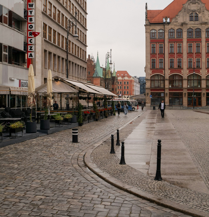 A cobblestone street leads toward historic buildings with a sidewalk cafe area under white umbrellas on the left.
