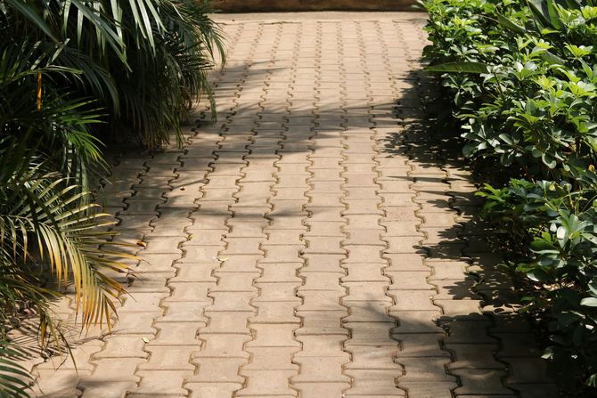 A stone pathway made of interlocking brown pavers, lined with green foliage and palm fronds on either side.